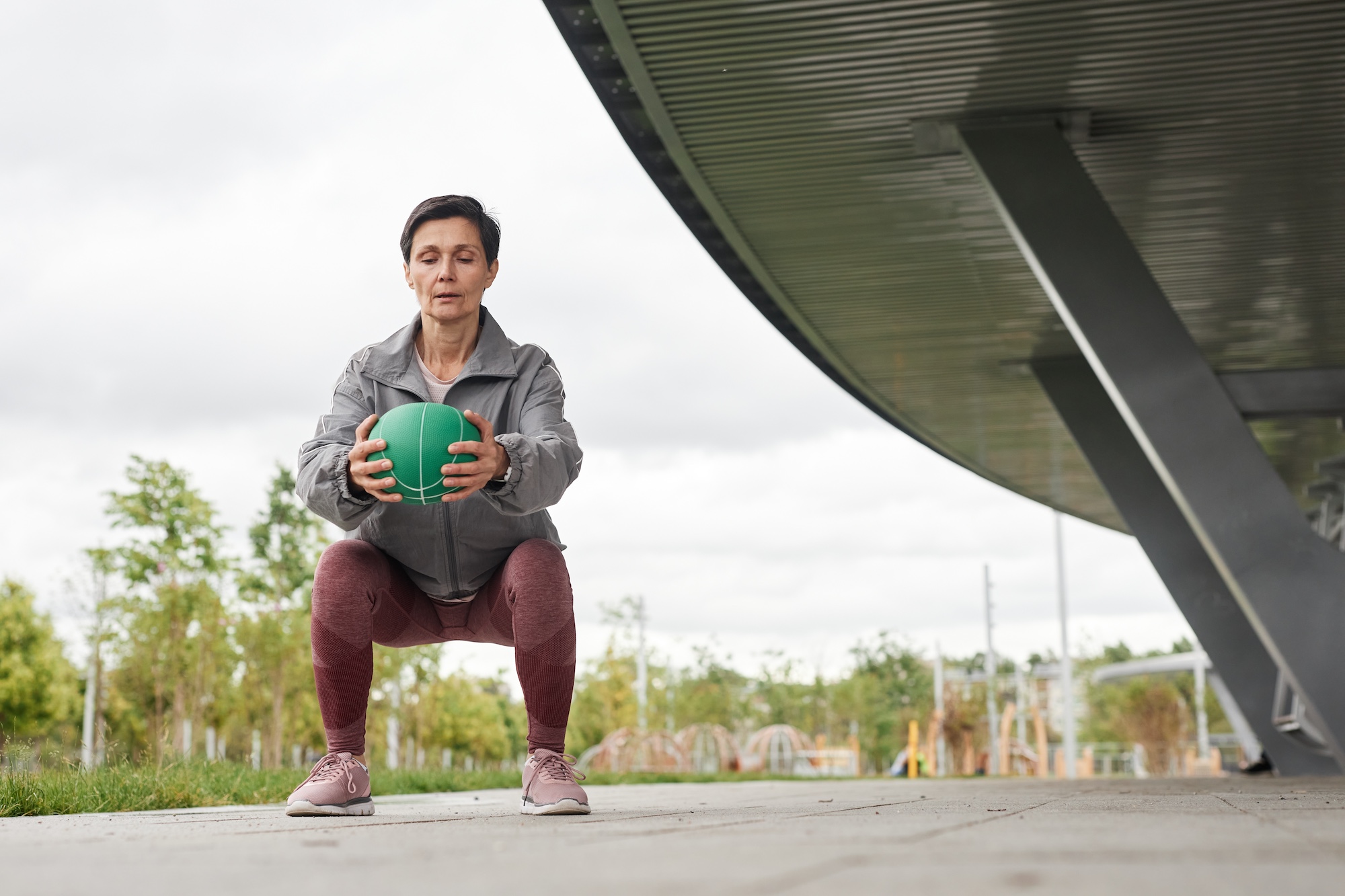 Elderly Woman Holding Ball in Squat Working out at City Park against Gray Sky